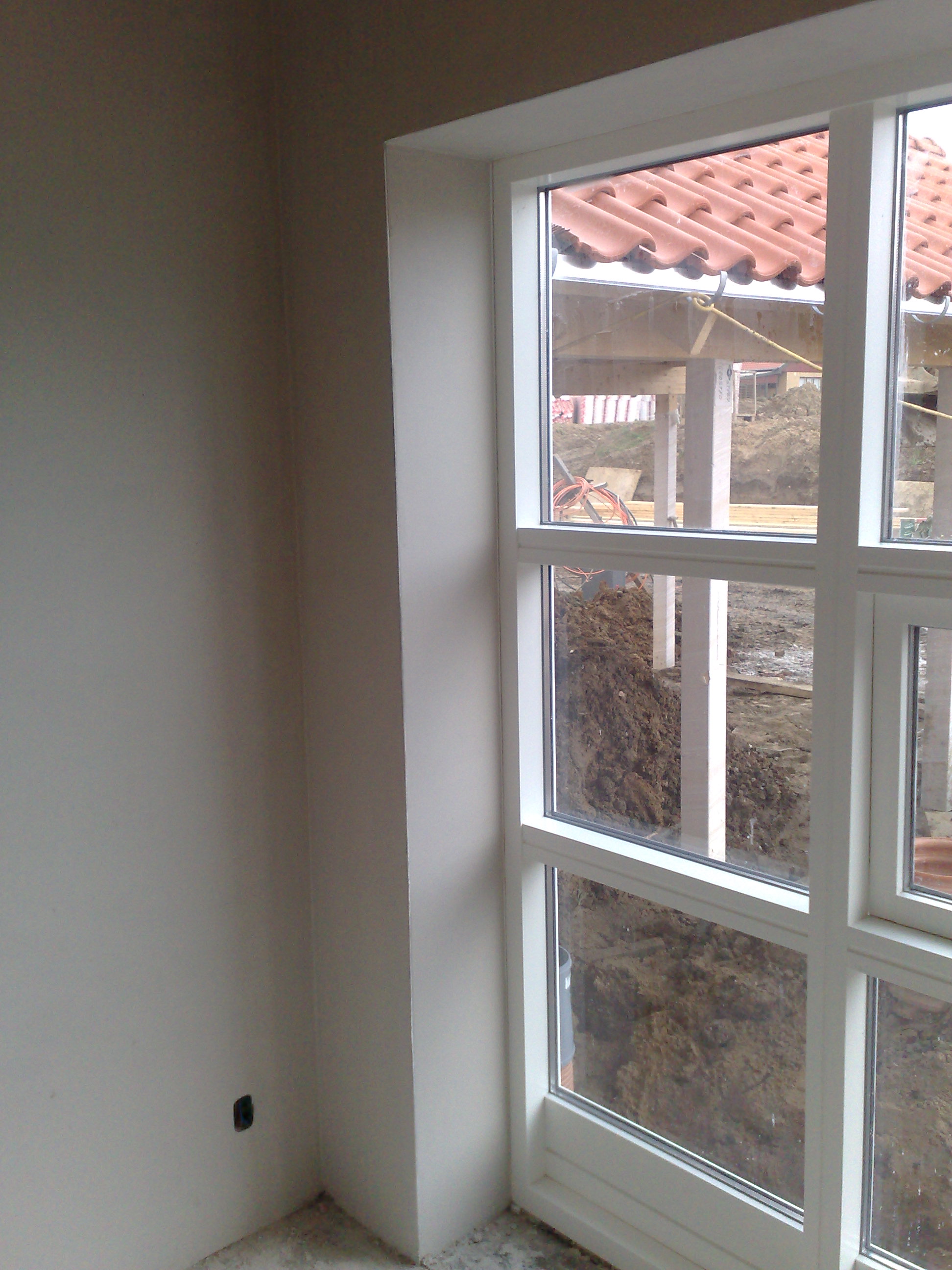 Wnętrze w trakcie prac wykończeniowych: gładka, jasnoszara ściana z widocznym gniazdkiem elektrycznym, białe obramowanie okna z widokiem na plac budowy z elementami konstrukcji dachu krytego...