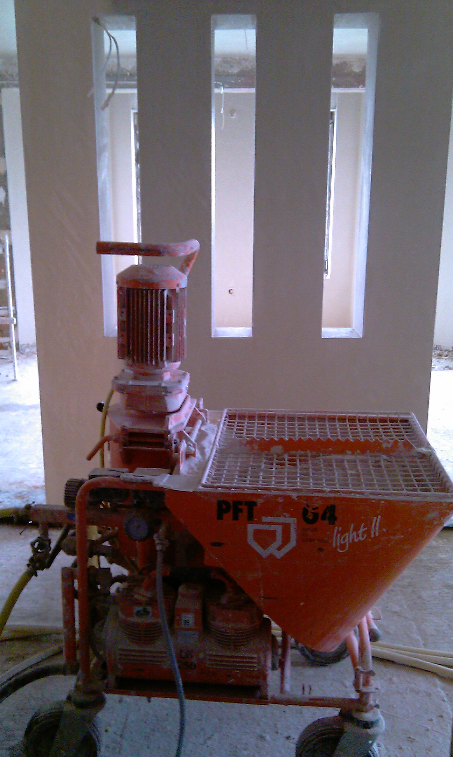 Pomarańczowa maszyna do wylewek gipsowych PFT G4 light II, z widocznymi śladami zaprawy, stoi na budowie na tle ściany z trzema pionowymi otworami okiennymi.