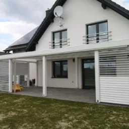 Nowoczesna, biała pergola z poziomymi lamelami, przylegająca do białego domu z czarnym dachem i panelami słonecznymi, widoczne dwa żółte krzesła wewnątrz konstrukcji.