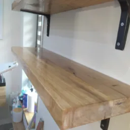 Dwie półki z drewna przymocowane do ściany za pomocą czarnych, metalowych wsporników. Na dolnej półce widoczne kosmetyki i przybory.