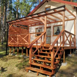 Wykończony drewniany taras z zadaszeniem przy domu szkieletowym, schody prowadzą na taras, widoczne las sosnowy w tle.