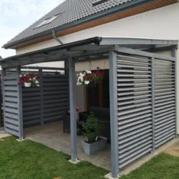 Szara, drewniana konstrukcja z żaluzjowymi ścianami przylegająca do jasnego domu z szarą dachówką, udekorowana wiszącymi kwiatami w doniczkach i roślinami w donicach na tarasie.