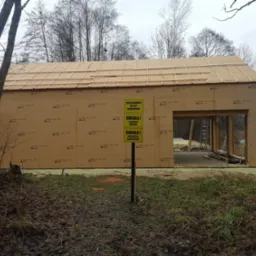 Surowa konstrukcja szkieletowa budynku w trakcie budowy, widoczne ściany z płyt OSB z nadrukiem, drewniana więźba dachowa, tablica ostrzegawcza 'Teren budowy wstęp zabroniony', w tle jezioro i drzewa.