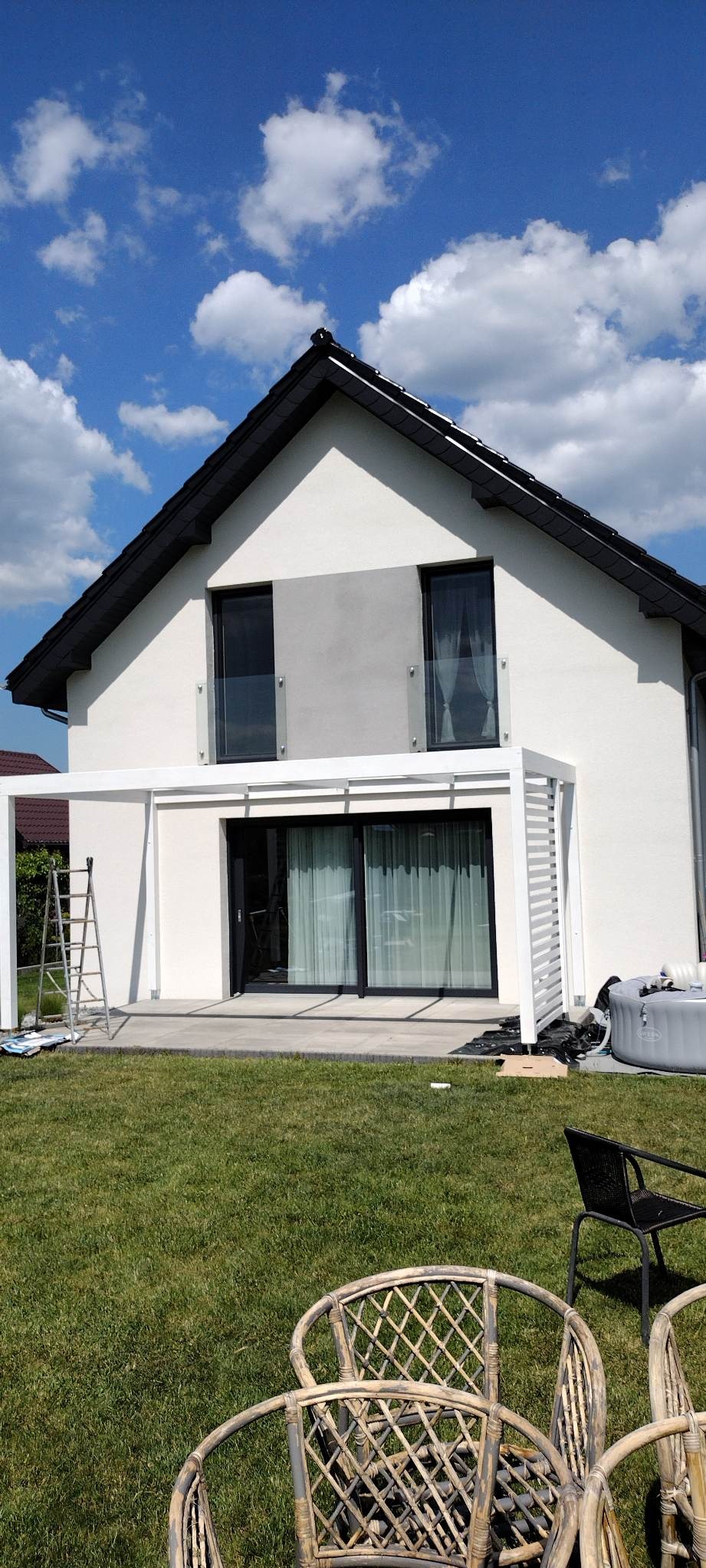 Nowoczesna, biała pergola przylegająca do domu z dużymi przesuwnymi drzwiami tarasowymi, drabina oparta o konstrukcję, trawnik, częściowo widoczne krzesła ogrodowe, jacuzzi w tle.