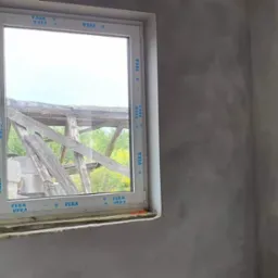 Ściana wewnętrzna po nałożeniu szarej warstwy wykończeniowej, widoczne okno z ramą VEKA, za oknem konstrukcja drewniana i zieleń.