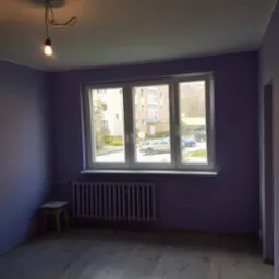 Pokój po malowaniu w Gdyni, ściany w odcieniach fioletu, białe okno z widokiem na budynek mieszkalny, drewniana podłoga, grzejnik pod oknem, surowa lampa z żarówką.