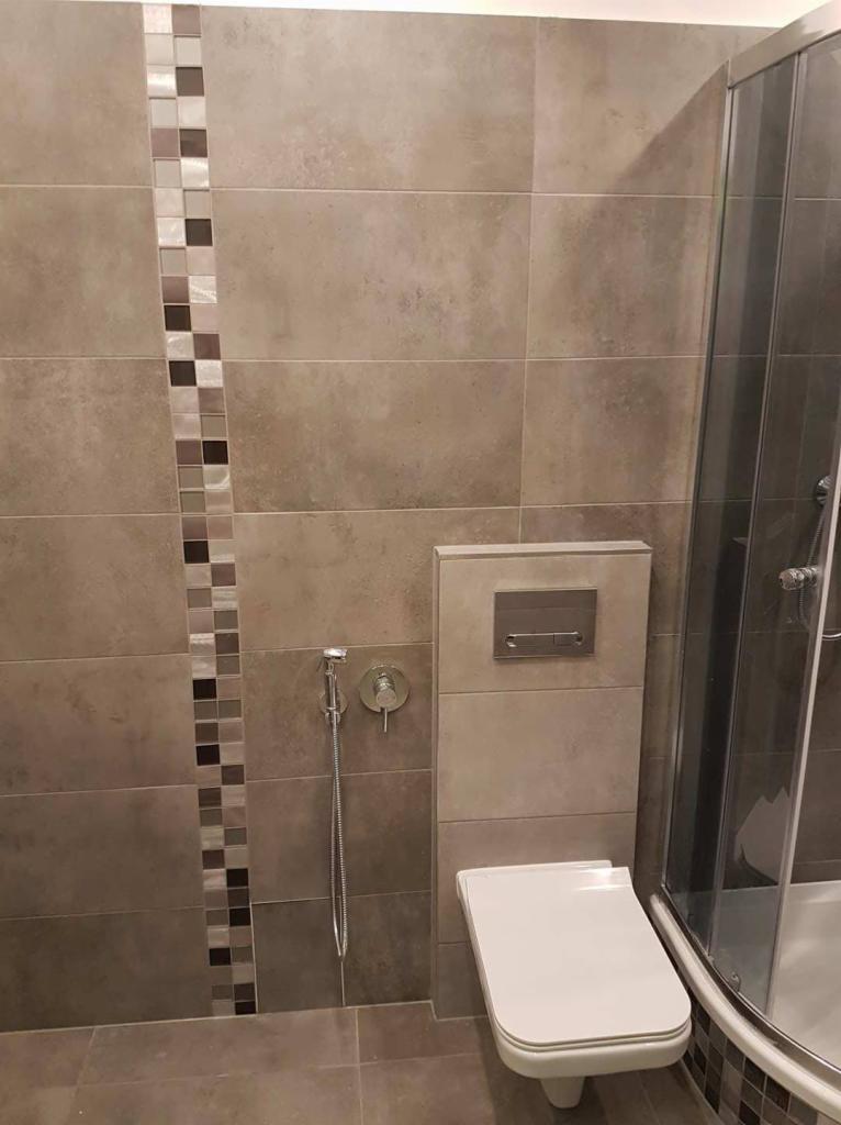 Nowoczesna łazienka z beżowymi płytkami, dekoracyjnym pasem mozaiki, wiszącą toaletą i chromowaną słuchawką bidetową przy ścianie.