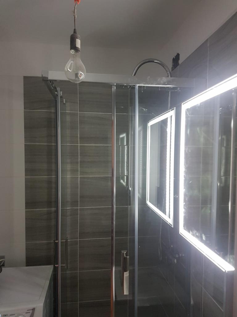 Szklana kabina prysznicowa z chromowanymi profilami i uchwytami, oświetlona żarówką na kablu, z podświetlanymi lustrami na ciemnych płytkach w tle.