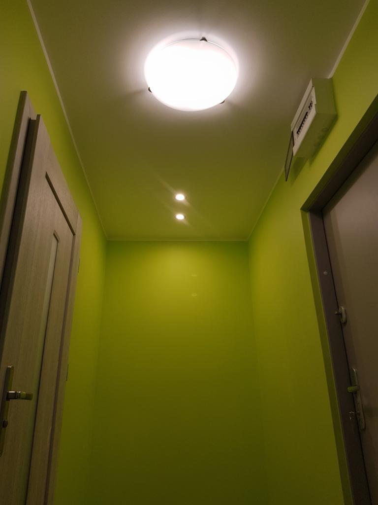 Korytarz pomalowany na limonkowy kolor z widocznymi drzwiami po bokach, oświetlony okrągłą lampą sufitową i dwoma punktowymi światłami.