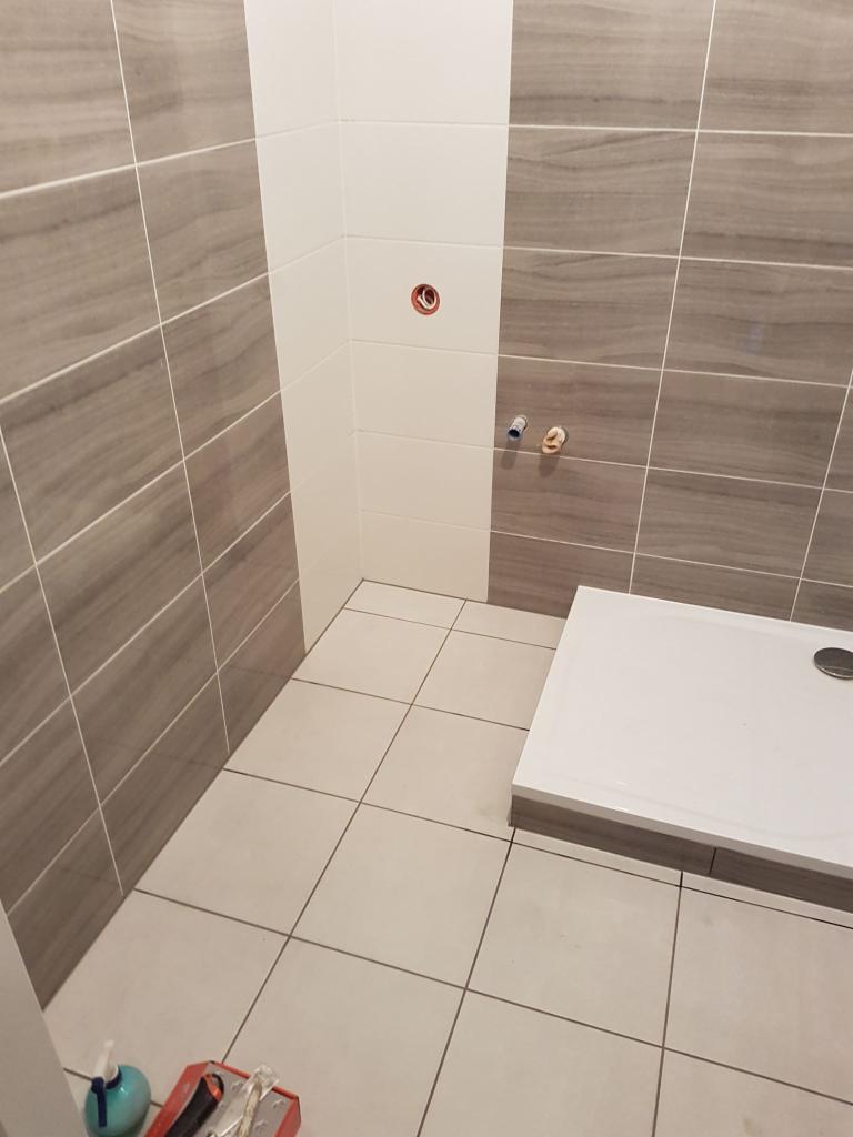 Nowoczesna łazienka w trakcie wykańczania: szare płytki imitujące drewno na ścianach, białe płytki na fragmencie ściany, biały brodzik z odpływem, narzędzia glazurnicze w rogu.