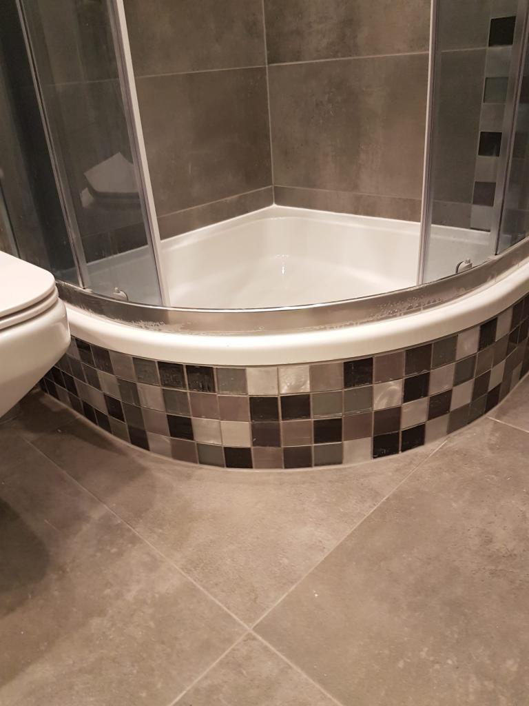 Szczegółowe ujęcie narożnej kabiny prysznicowej z przesuwnymi drzwiami, wykończonej mozaiką w odcieniach szarości i czerni, z widocznym fragmentem toalety po lewej stronie i szarą, kwadratową podłogą.