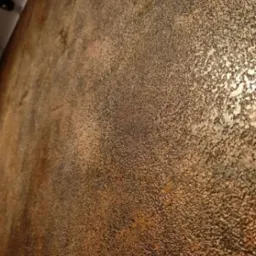 Tekstura ściany z efektem rdzy, nier&oacute;wnomierne wykończenie z połyskującymi fragmentami, widoczne detale faktury na brązowo-złotym tle.