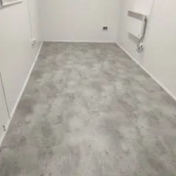 Szara posadzka mikrocementowa w pustym, jasnym pomieszczeniu z białymi ścianami i grzejnikiem elektrycznym.