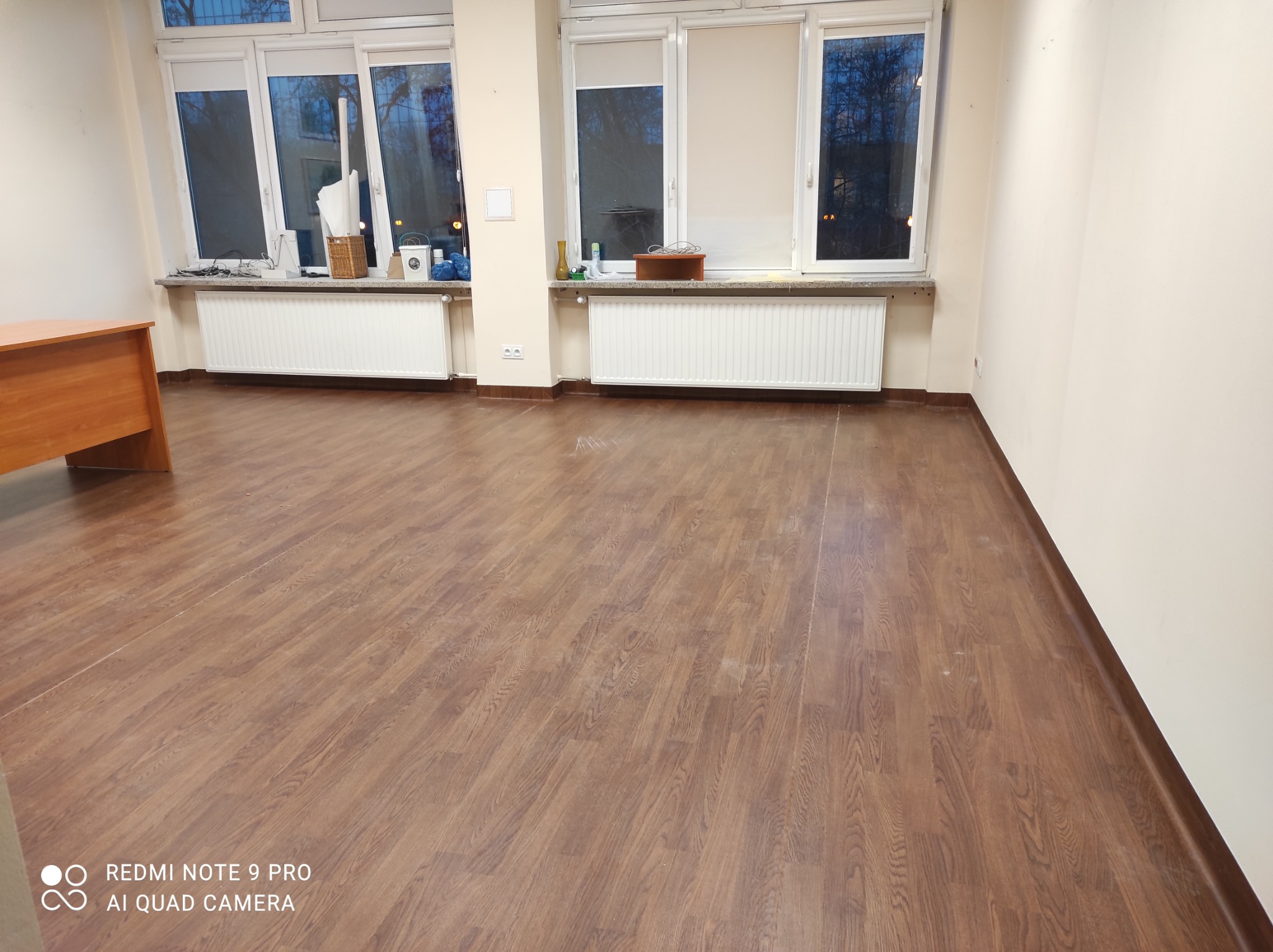 Brązowa podłoga imitująca drewno w pustym, jasnym pomieszczeniu biurowym z białymi ścianami, grzejnikami pod oknami i biurkiem w rogu.