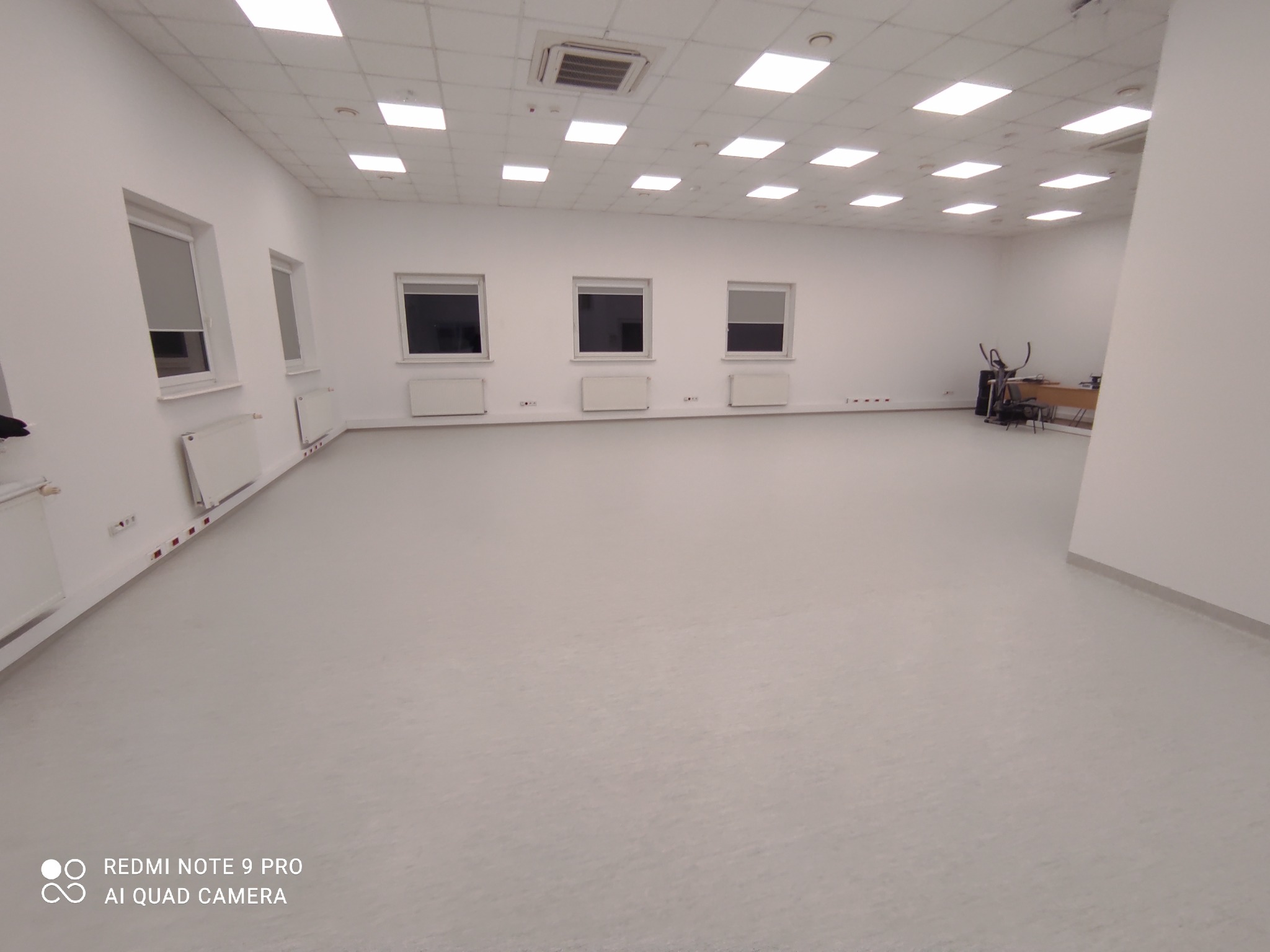 Jasne, przestronne biuro po remoncie, z nową, jednolitą posadzką w odcieniach szarości, widoczne okna z roletami, grzejniki oraz sprzęt biurowy w oddali.