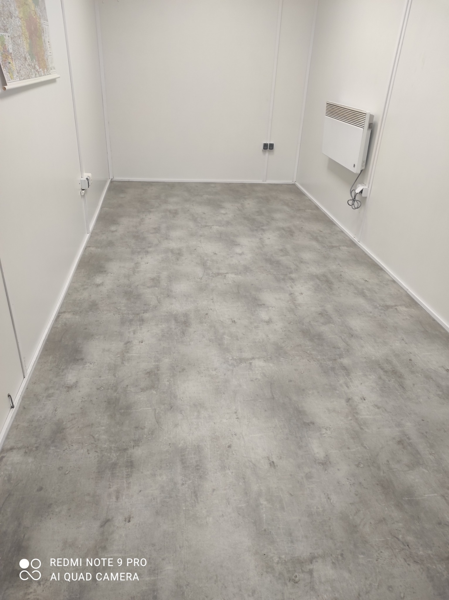 Szara posadzka mikrocementowa w pustym, jasnym pomieszczeniu z białymi ścianami i grzejnikiem elektrycznym.