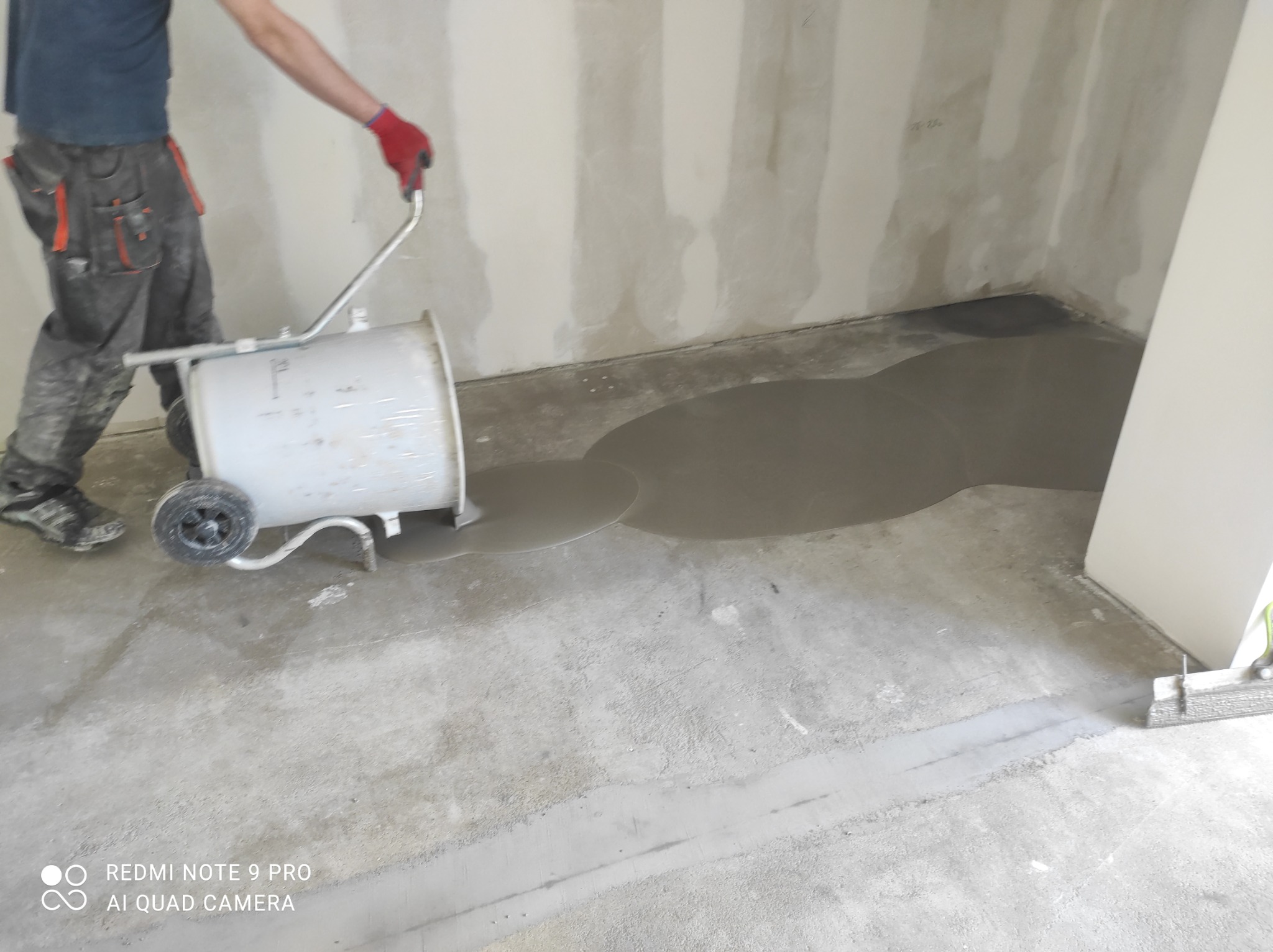 Aplikacja wylewki samopoziomującej w pomieszczeniu, widoczny pracownik w roboczym ubraniu i czerwonych rękawicach, używający wózka z pojemnikiem do rozprowadzania masy.