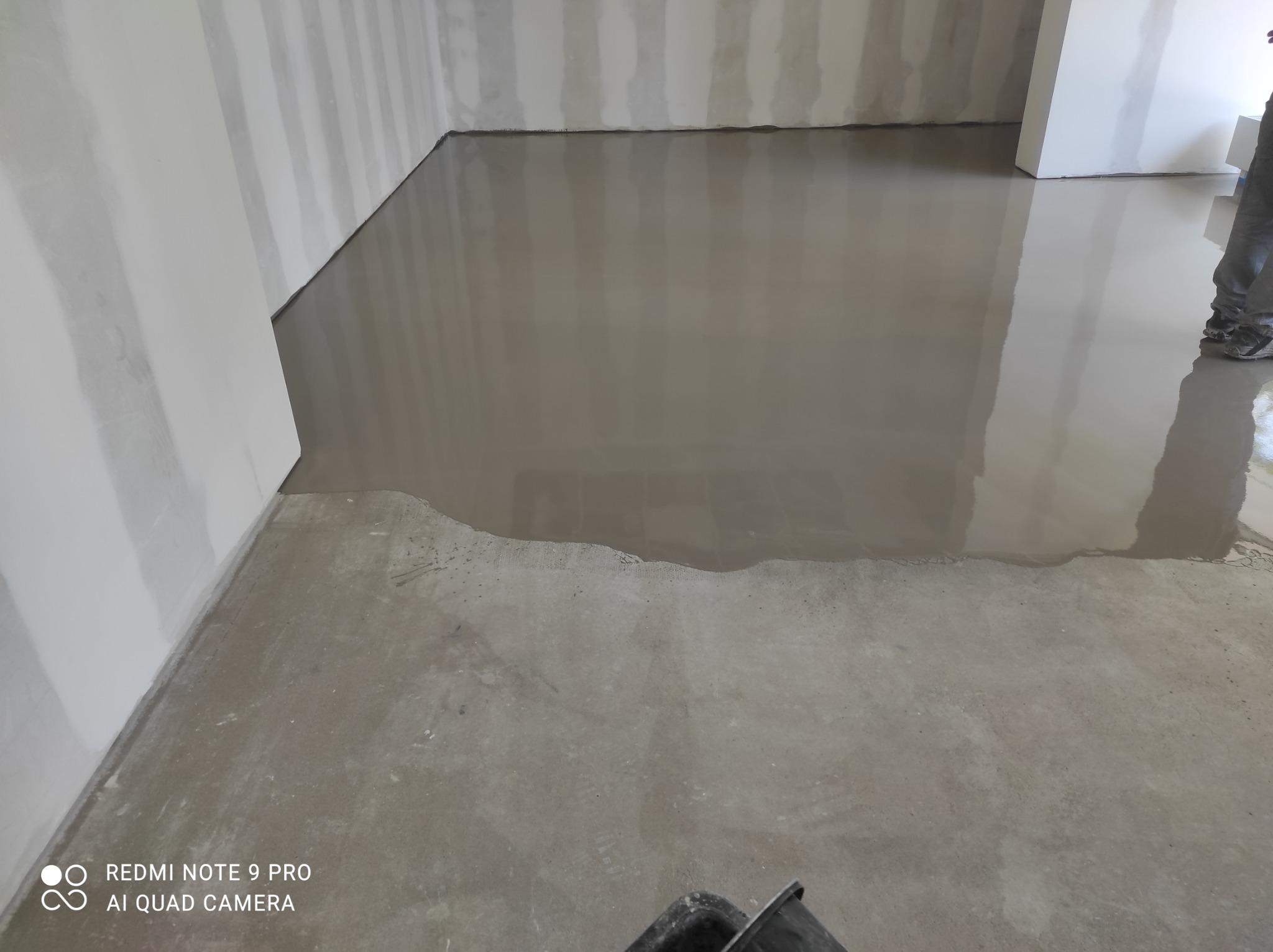 Wylewka samopoziomująca, świeżo rozprowadzona, z widocznymi śladami narzędzi i fragmentem osoby w roboczych spodniach, na tle przygotowanych ścian.
