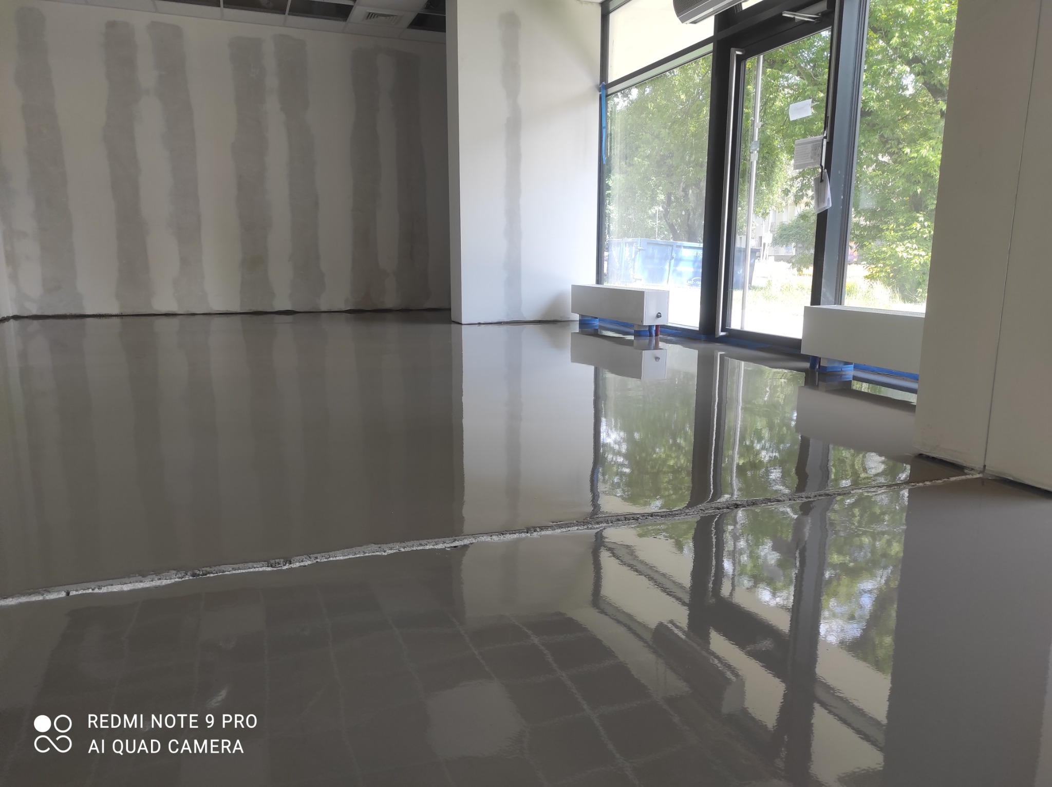 Świeżo wylana, gładka, szara posadzka w jasnym pomieszczeniu biurowym, widoczne ślady po kratownicy, duże okna z widokiem na zieleń na zewnątrz.