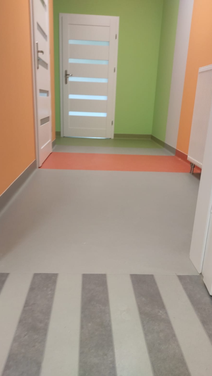 Korytarz z szarą posadzką w paski, pomarańczowymi i zielonymi ścianami, oraz białymi drzwiami z przeszkleniami.