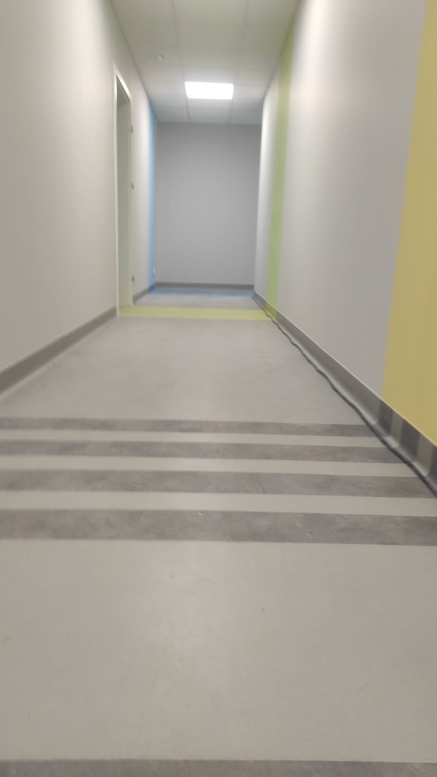 Korytarz biurowy z szarą posadzką w pasy i kolorowymi akcentami na ścianach, widok z perspektywy podłogi w stronę końca korytarza.