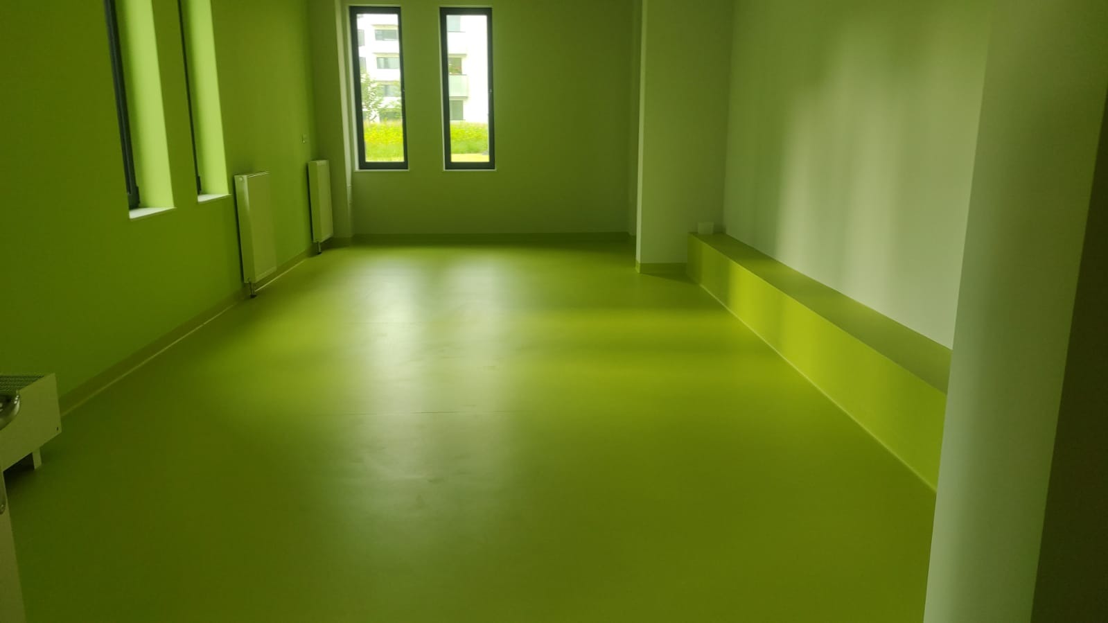 Jasnozielona, jednolita posadzka w pustym pomieszczeniu biurowym, z widocznymi grzejnikami pod oknami i wbudowaną ławką wzdłuż ściany.