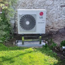 Biała pompa ciepła LG Therma V zamontowana na chromowanym stelażu przed ścianą budynku z jasnym, łuszczącym się tynkiem, otoczona zielenią trawnika i krzewów.