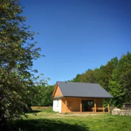 Niewielki, parterowy dom z drewnianą elewacją i szarym dachem na tle błękitnego nieba i zieleni drzew.  Zadaszony taras z drewnianymi ławkami. Część ściany pomalowana na biało.