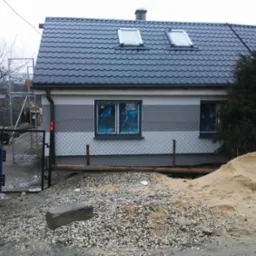 Dom jednorodzinny z nową elewacją w odcieniach szarości, okna zabezpieczone folią, widoczny kontener na gruz, rusztowanie i pryzma piasku.
