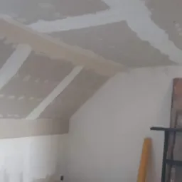 Poddasze w trakcie wykańczania: płyty gipsowo-kartonowe na skosach i suficie, z widocznymi łączeniami szpachlowanymi na biało, fragment otworu w suficie, drabina i materiały budowlane w tle.