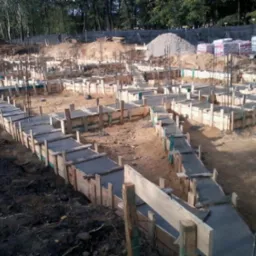 Przygotowane szalunki i zbrojenie pod wylewkę fundamentów na terenie budowy, widoczne stosy materiałów budowlanych w tle.