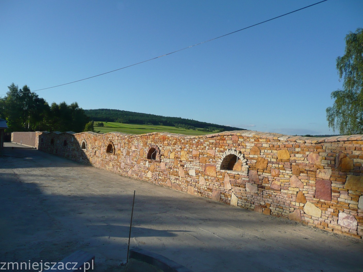 Długi mur z piaskowca z półokrągłymi wnękami, prowadzący wzdłuż drogi, w tle zielone wzgórza i błękitne niebo.