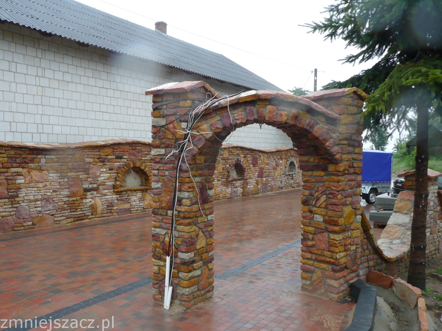 Kamienna brama wejściowa z łukiem, wykonana z piaskowca w odcieniach brązu i pomarańczy, z widocznymi kablami na jednym z filarów, prowadząca na wybrukowany dziedziniec z kamiennymi murkami i wnękami.