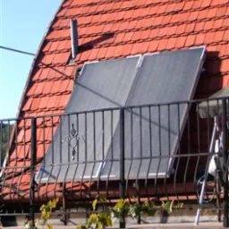 Dwa kolektory słoneczne Vitosol 100F zamontowane na czerwonym dachu z dachówki, widoczne zza ozdobnej, metalowej balustrady balkonu z zielonymi pnączami.