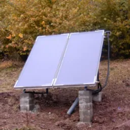 Dwa panele solarne zamontowane na betonowych bloczkach, z widocznymi rurami, na tle jesiennego krajobrazu z brązowymi i żółtymi liśćmi.