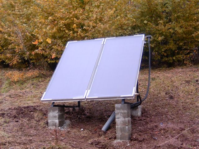 Dwa panele solarne zamontowane na betonowych bloczkach, z widocznymi rurami, na tle jesiennego krajobrazu z brązowymi i żółtymi liśćmi.