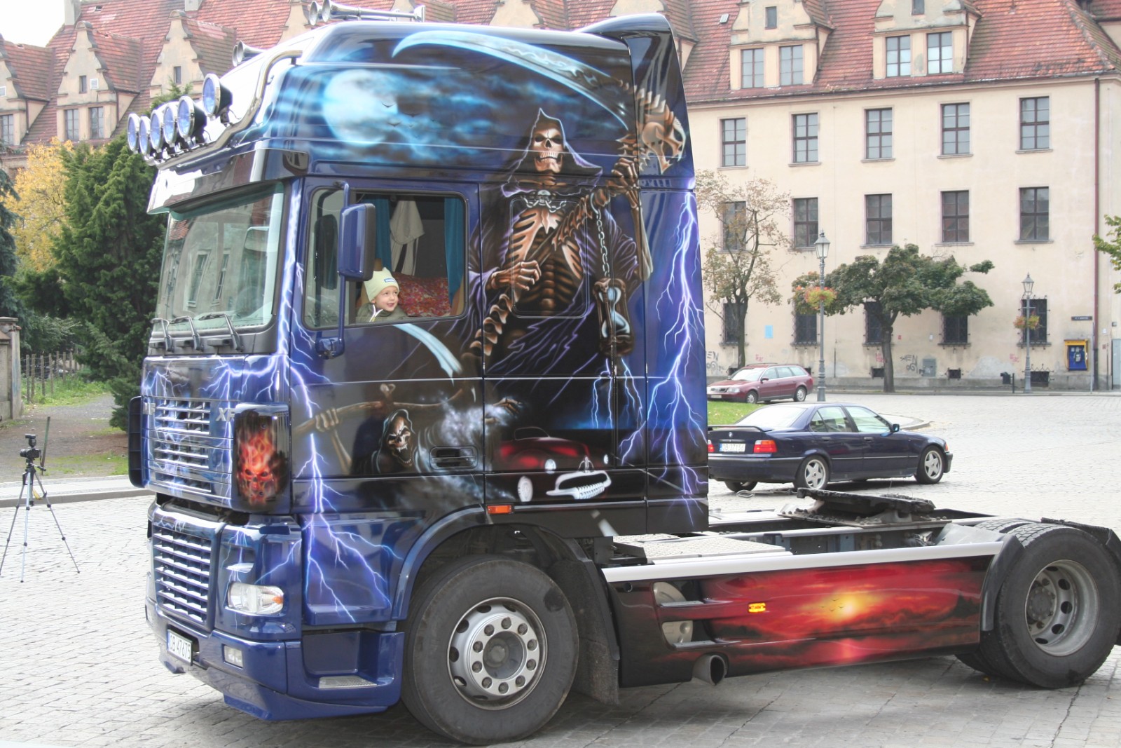 Ciągnik siodłowy z efektownym, artystycznym lakierowaniem airbrush przedstawiającym postacie śmierci i motywy błyskawic; w kabinie widoczne dziecko w czapce.