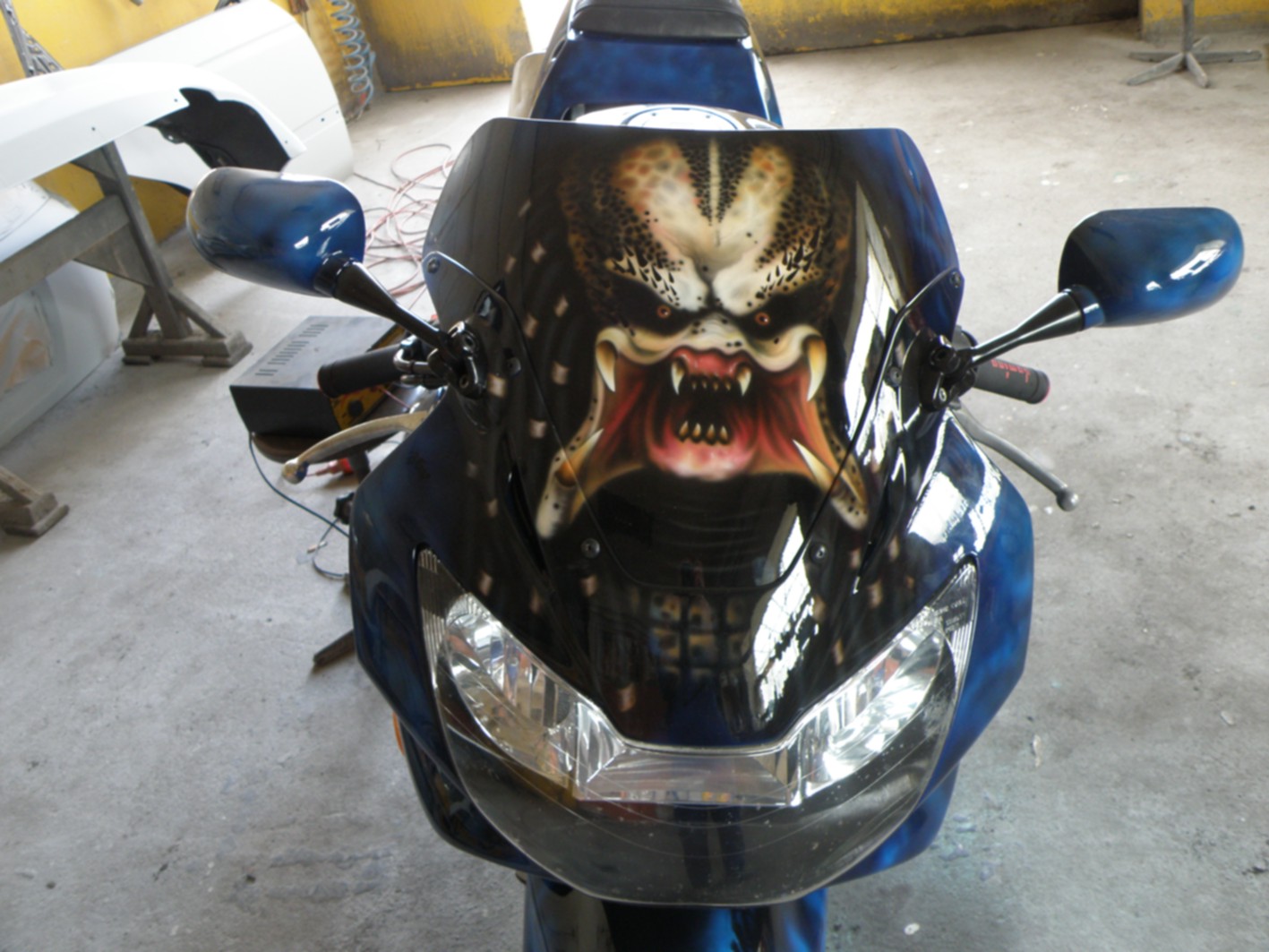 Motocykl z wizualnym tuningiem: aerograf przedstawiający postać Predatora na przedniej owiewce, niebieskie detale lakiernicze, widoczne elementy karoserii w tle.