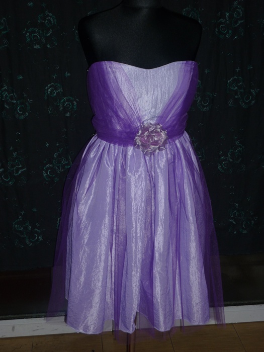 Liliowa sukienka bez ramiączek z tiulowym pasem i kwiatową aplikacją, prezentowana na manekinie na tle ciemnej tkaniny z delikatnym wzorem.