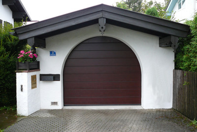 Garaż z brązową bramą segmentową w kształcie łuku, białe ściany z dekoracyjnymi drewnianymi elementami, skrzynka na listy i donica z kwiatami.