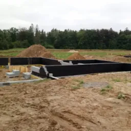 Budowa fundamentów domu z bloczków betonowych, widoczne izolacja pionowa, szalunki, zaplecze budowy i hałdy ziemi w tle.