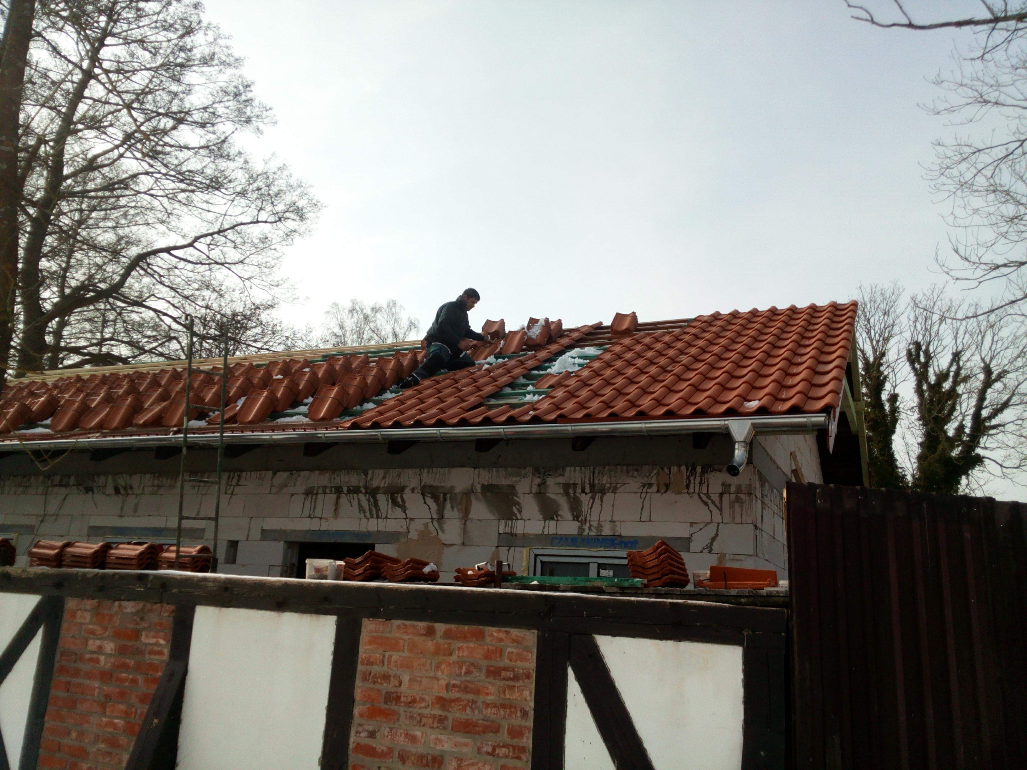 Dekarz układa czerwoną dachówkę na częściowo ośnieżonym dachu domu w trakcie budowy, widoczne drzewa bez liści w tle i fragment muru pruskiego.