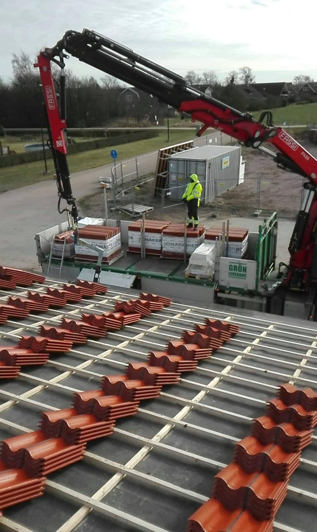 Układanie czerwonej dachówki karpiówki na łatach dachowych z użyciem dźwigu Fassi i platformy z paletami dachówek; pracownik w żółtej kamizelce nadzoruje proces.