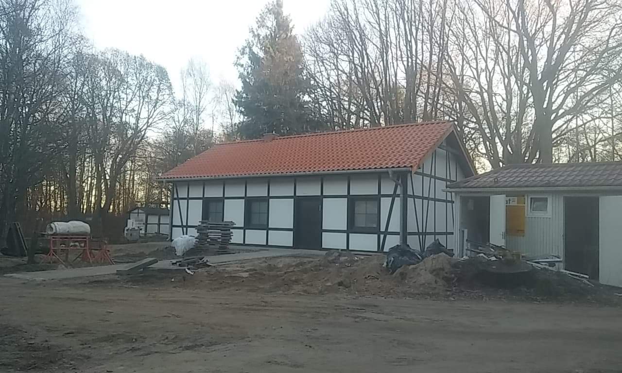 Budynek w stylu szachulcowym z czerwoną dachówką, widoczne materiały budowlane i otoczenie leśne.