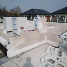 Wznoszenie ścian parteru domu z bloczków Solbet, widoczne narzędzia murarskie i zapas materiału budowlanego w foliowych opakowaniach, w tle zabudowa jednorodzinna.