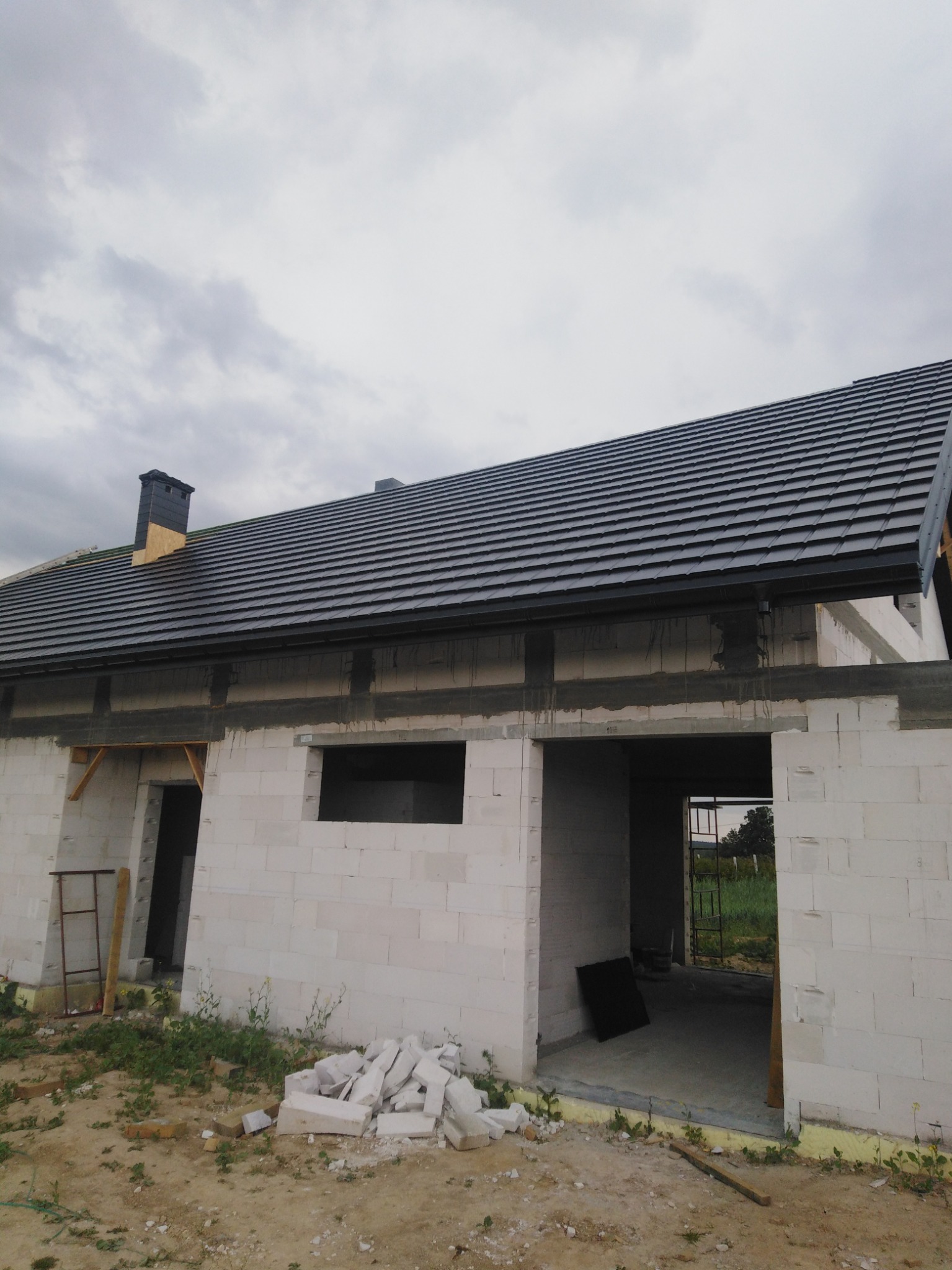 Surowy stan domu z białego pustaka z ciemną dachówką, widoczny komin i fragment nieba.