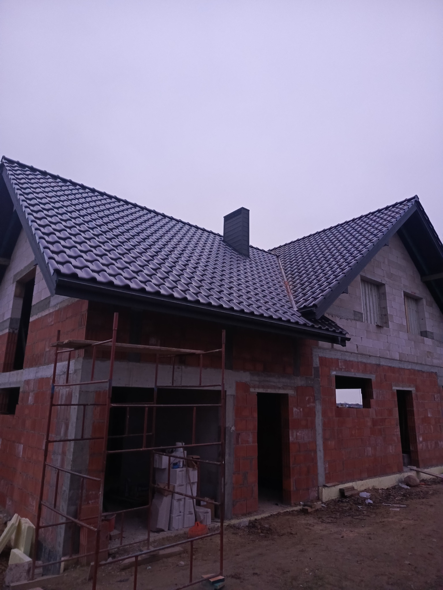 Niedokończony dom z ciemną dachówką, widoczne rusztowania i ceglane ściany w surowym stanie. Dach pokryty, okna jeszcze nie wstawione. Pochmurne niebo w tle.
