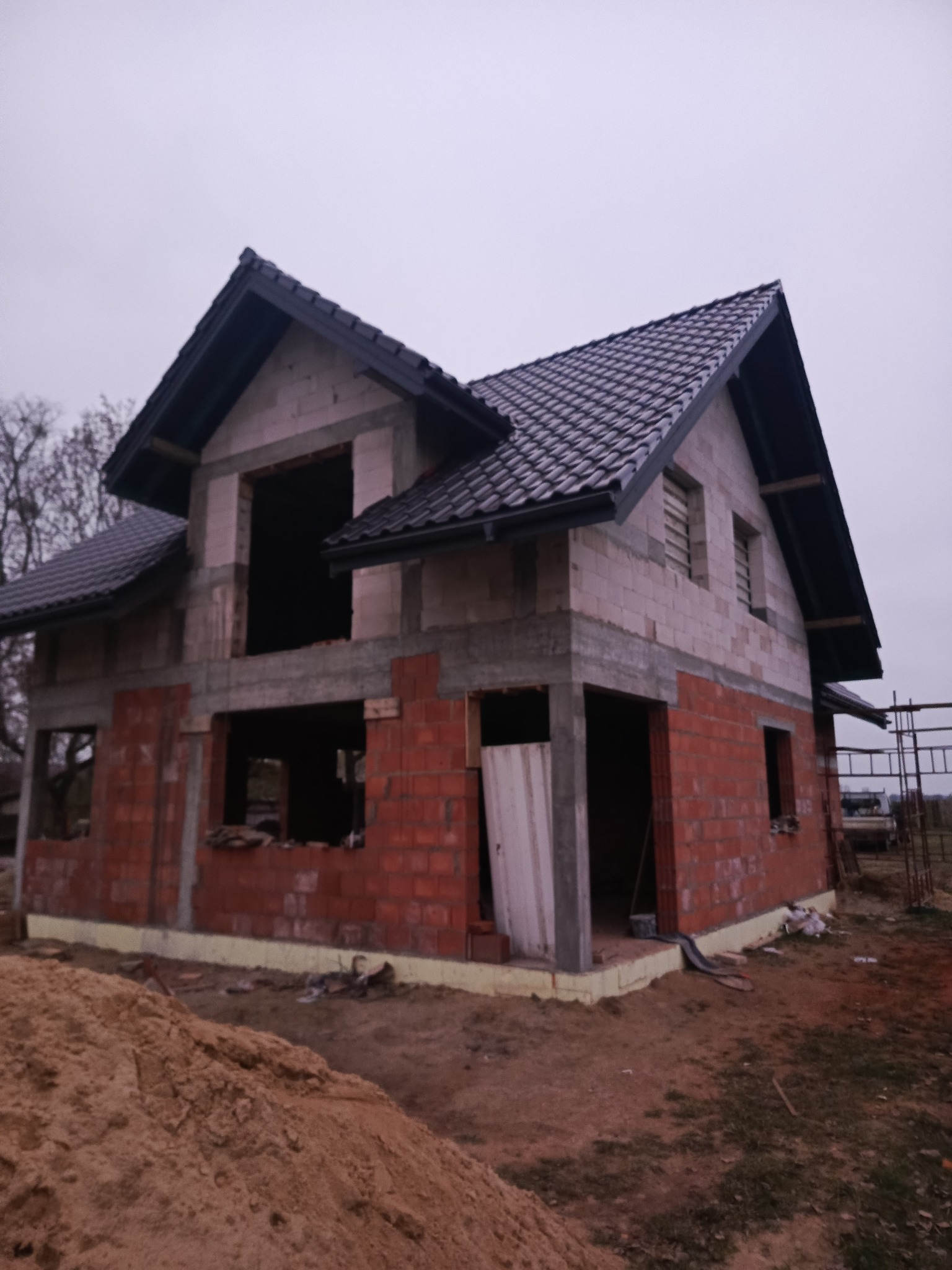 Dwukondygnacyjny dom w trakcie budowy: czerwona cegła na dole, szare bloczki na górze, czarny dach z dachówki. Widoczne otwory okienne i drzwiowe, teren budowy.