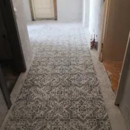 Wnętrze w trakcie prac wykończeniowych: wzorzysta podłoga ułożona na świeżym podkładzie, widoczne tuby z klejem i fragment starej podłogi w przejściu.