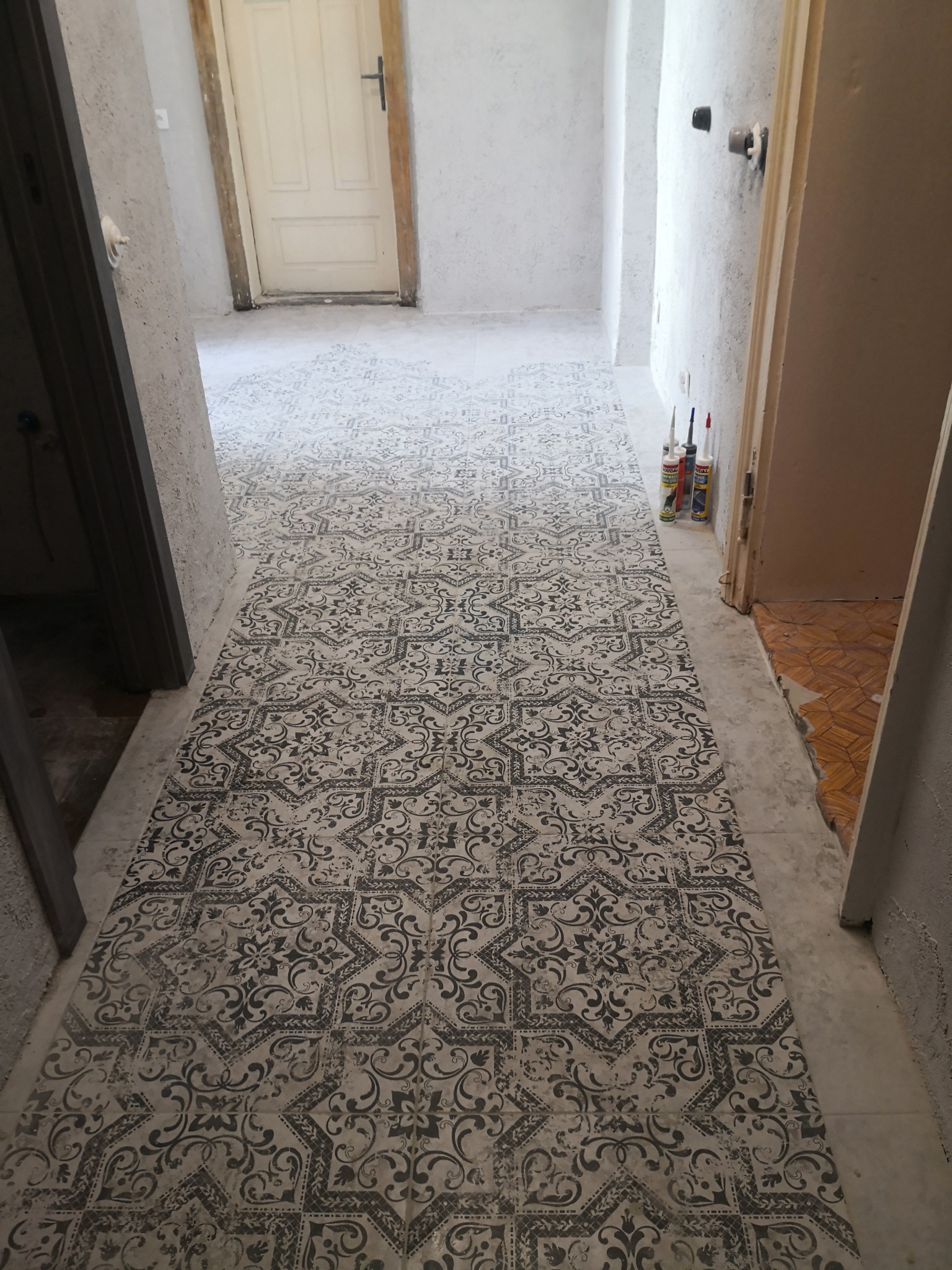Wnętrze w trakcie prac wykończeniowych: wzorzysta podłoga ułożona na świeżym podkładzie, widoczne tuby z klejem i fragment starej podłogi w przejściu.